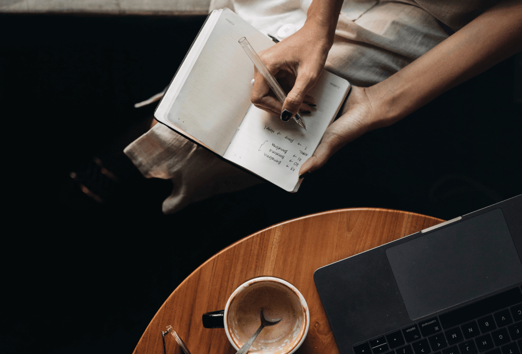 A woman's hand writes a note in a notebook, there is a cup of coffee on the table. Top view.