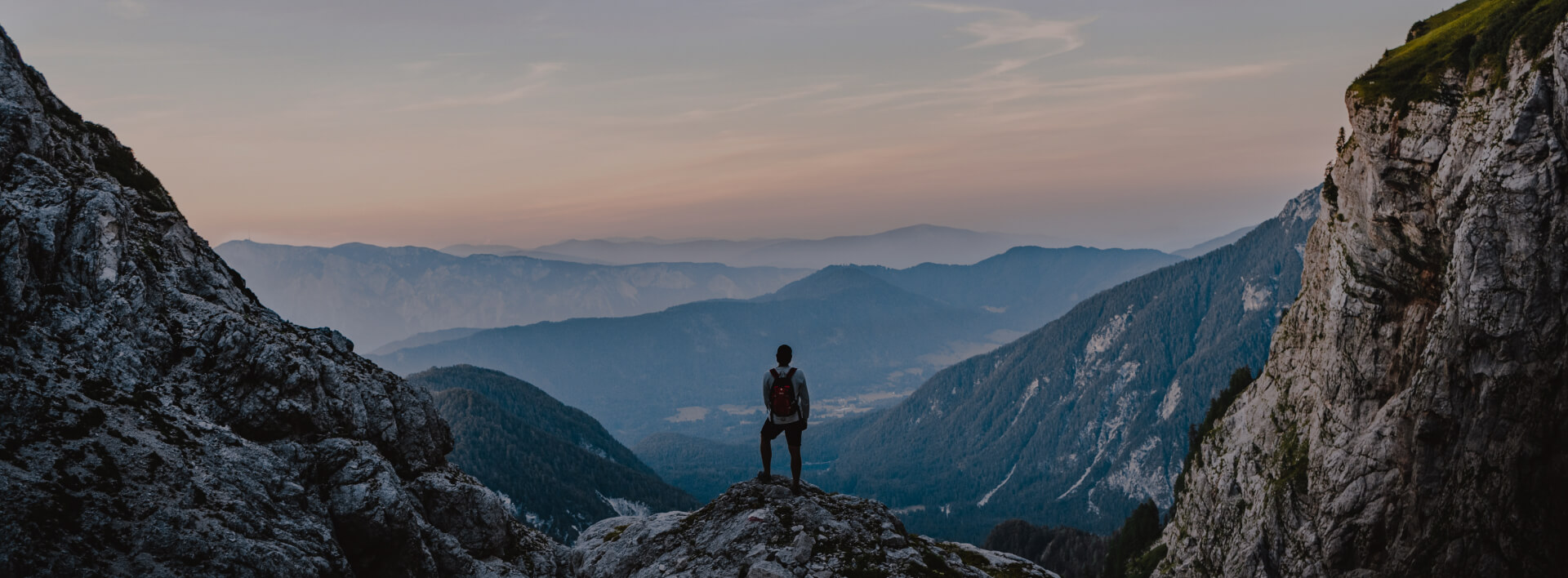 Silhouette of a man standing on top of a cliff, with high mountain ranges in the background.