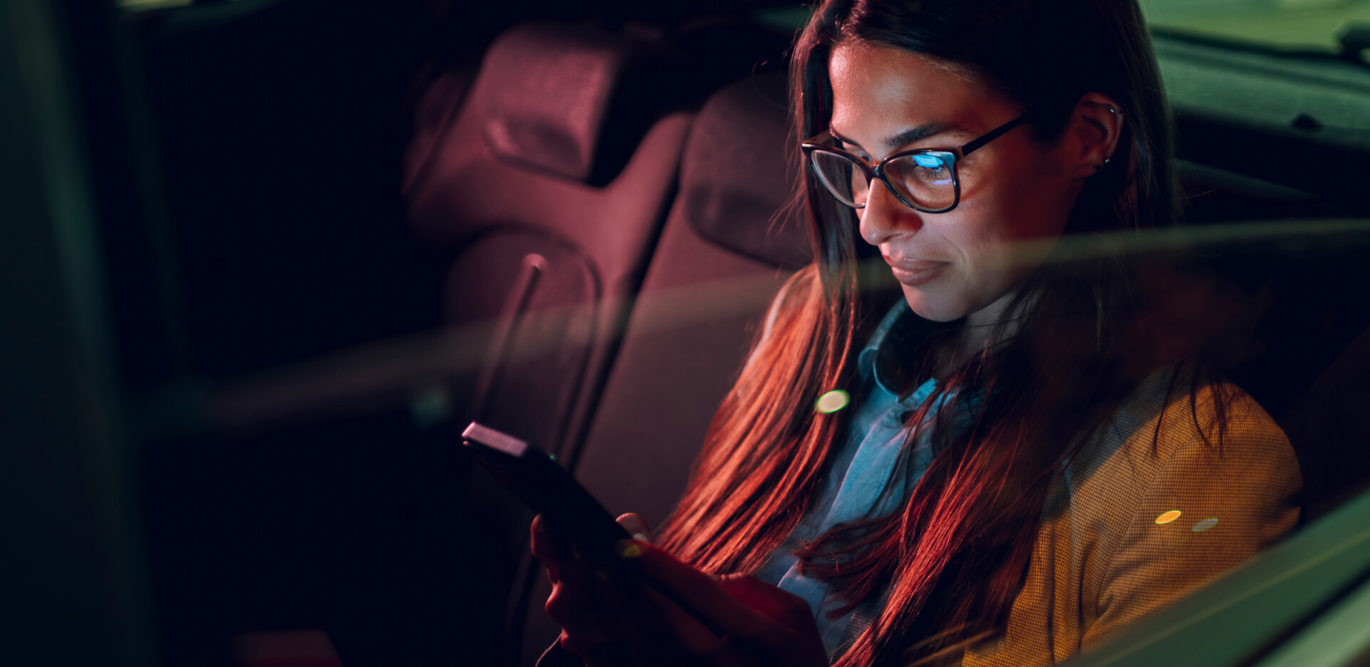 Girl with glasses in the back seat of a car looking at the phone screen.